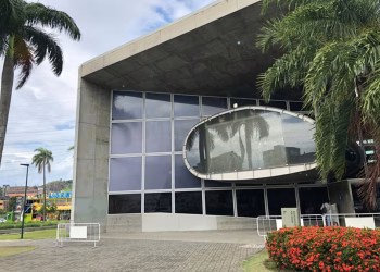 Fachada de uma das Naves do Conhecimento no Rio de Janeiro, prédio de concreto com grande janela oval espelhada e palmeiras na entrada