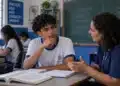 Estudante do ensino médio conversa com orientadora em sala de aula, com livros e cadernos sobre a mesa.