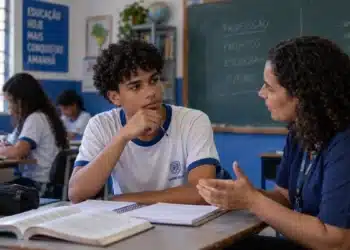 Estudante do ensino médio conversa com orientadora em sala de aula, com livros e cadernos sobre a mesa.
