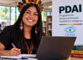 Imagem ilustrativa de estudante indígena com cocar de penas e pintura facial, sorrindo enquanto faz anotações ao lado de um notebook em uma biblioteca, com banner do PDAI e da Capes ao fundo