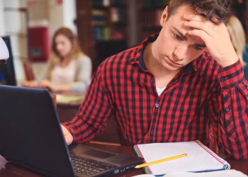 Imagem ilustrativa de estudante preocupado, com a mão na testa diante de um notebook e de um caderno em uma biblioteca, em referência à depressão em estudantes de Medicina