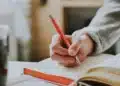 Pessoa escreve com caneta vermelha em caderno, revisando a grafia de fim de semana e outras locuções sem hífen