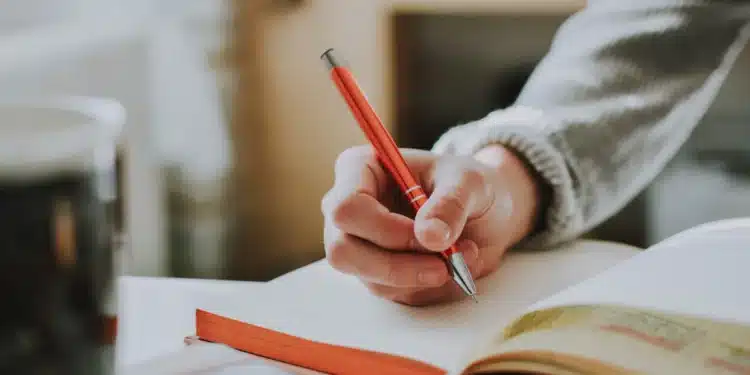 Pessoa escreve com caneta vermelha em caderno, revisando a grafia de fim de semana e outras locuções sem hífen