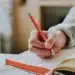 Pessoa escreve com caneta vermelha em caderno, revisando a grafia de fim de semana e outras locuções sem hífen