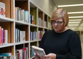 Bibliotecária segura livro entre estantes; a profissão de bibliotecário exige bacharelado e registro no CRB