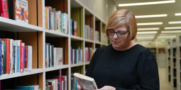 Bibliotecária segura livro entre estantes; a profissão de bibliotecário exige bacharelado e registro no CRB