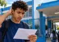 Estudante do Enem 2026 observa documento com preocupação diante de uma escola, representando as dúvidas sobre a possibilidade de alterar o local de prova devido à distância.