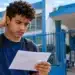 Estudante do Enem 2026 observa documento com preocupação diante de uma escola, representando as dúvidas sobre a possibilidade de alterar o local de prova devido à distância.