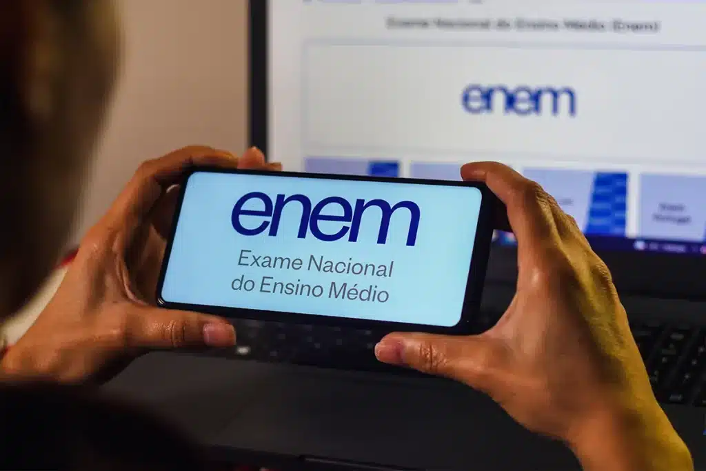 Pessoa segura celular com identificação do Enem diante de notebook, representando a utilização das notas em processos seletivos.
