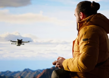 Imagem ilustrativa de piloto de jaqueta amarela segurando o controle enquanto acompanha com os olhos um drone em voo sobre montanhas, representando quem precisa fazer a prova da Anac para drones
