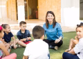 Imagem ilustrativa de profissional sentada no gramado em roda de conversa com crianças no pátio da escola, em referência à atuação de psicólogo e assistente social nas escolas públicas