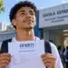 Estudante brasileiro segura comprovante de inscrição do Enem 2026 em frente a uma escola pública, com mochila nas costas.