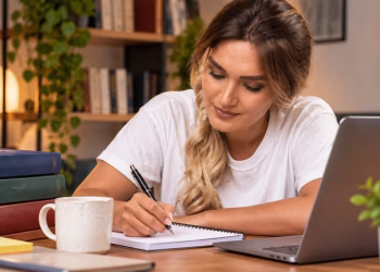 Imagem ilustrativa de mulher fazendo anotações em caderno ao lado de livros e notebook, em referência à dúvida sobre se a especialização exige trabalho de conclusão de curso