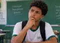 Estudante de escola pública com expressão pensativa e mão apoiada no queixo, em sala de aula, ilustrando dúvidas sobre o pagamento do Pé-de-Meia.