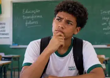 Estudante de escola pública com expressão pensativa e mão apoiada no queixo, em sala de aula, ilustrando dúvidas sobre o pagamento do Pé-de-Meia.