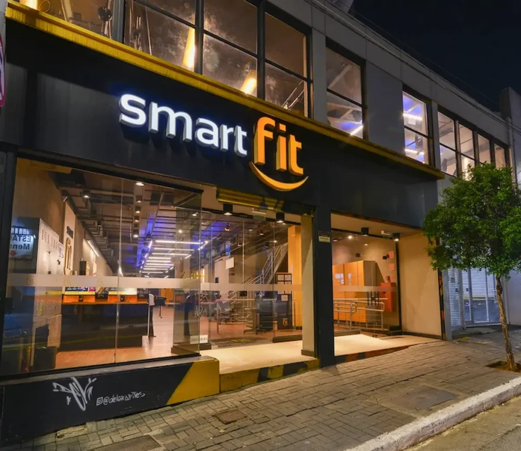 Fachada de uma academia Smart Fit à noite, com letreiro iluminado e entrada de vidro.