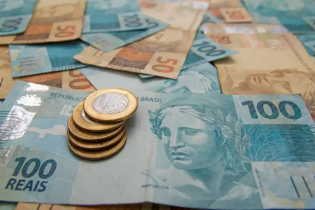 Cédulas de R$ 100 e R$ 50 com moedas de R$ 1 empilhadas, representando o incentivo Enem de R$ 200 do Pé-de-Meia.