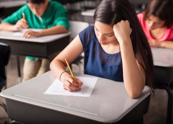 Estudante de blusa azul responde a uma prova com lápis em sala de aula, com outros alunos ao fundo