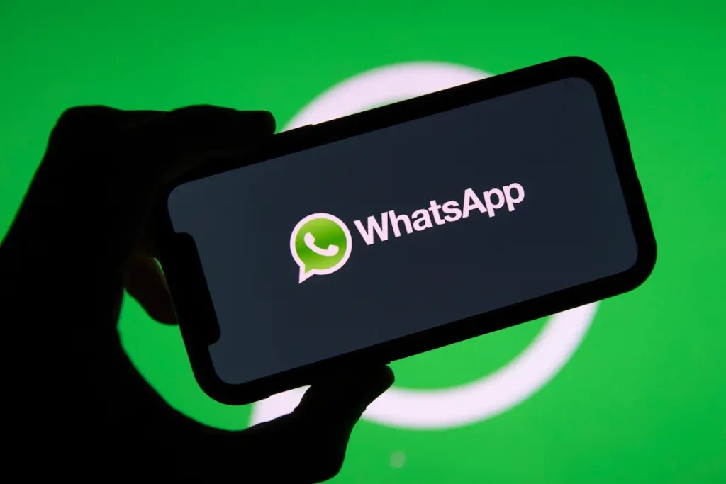 Mão segurando um celular na horizontal com o nome e o logotipo do WhatsApp na tela, sobre fundo verde.